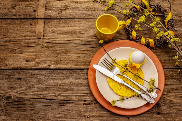 Easter table setting on old wooden background. Spring holiday card template. Cutlery, crochet napkin, egg, flowering sprigs of dogwood, cup, wreath