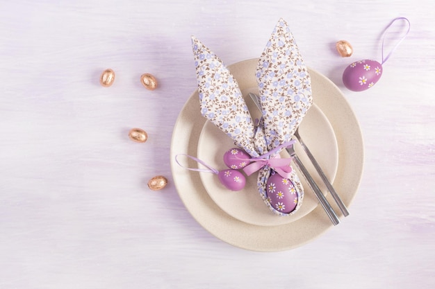 Easter table setting White plate with a napkin folded in the shape of a rabbit Easter and chocolate eggs on a pink background Happy Easter holiday concept Top view flat lay