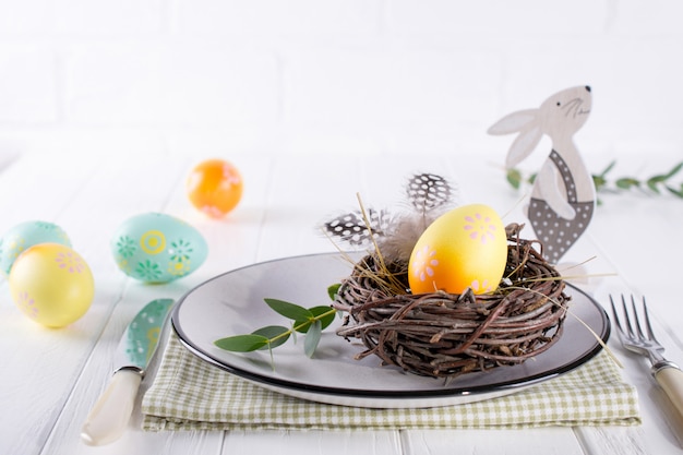 Easter table setting with  white plate, textile napkin yellow decorative chicken egg in the nest, mimosa flowers, feathers and spring easter decoration on white table