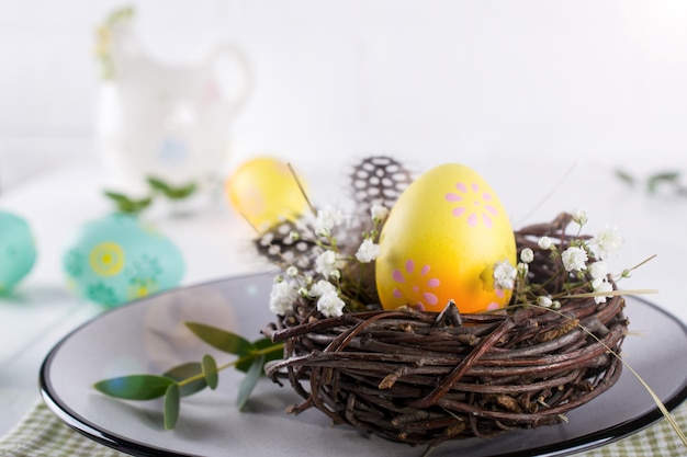 Easter table setting with  white plate, textile napkin yellow decorative chicken egg in the nest, mimosa flowers, feathers and spring easter decoration on white table