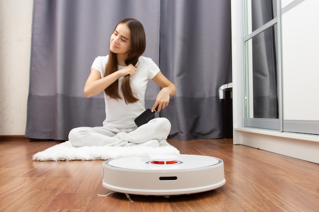 Easy cleaningYoung European girl combs her hair while robot vacuum cleaner collects dust