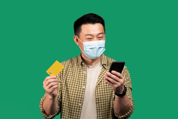 Easy payments asian middle aged man in face mask using cellphone and credit card buying goods online