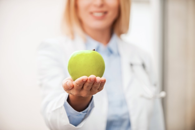 Eat healthy food! Close-up of female doctor in white uniform holding green apple and smiling while leaning at the glass wall