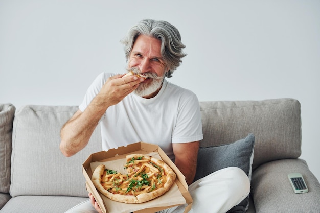 Eats delicious pizza while watching TV show Senior stylish modern man with grey hair and beard indoors