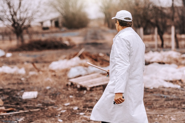 Ecologist in white uniform and cap on head holding clipboard while walking on landfill.