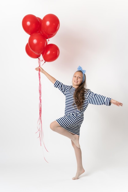 Ecstatic active girl holding bunch of red balloons in hand standing on one leg on white background