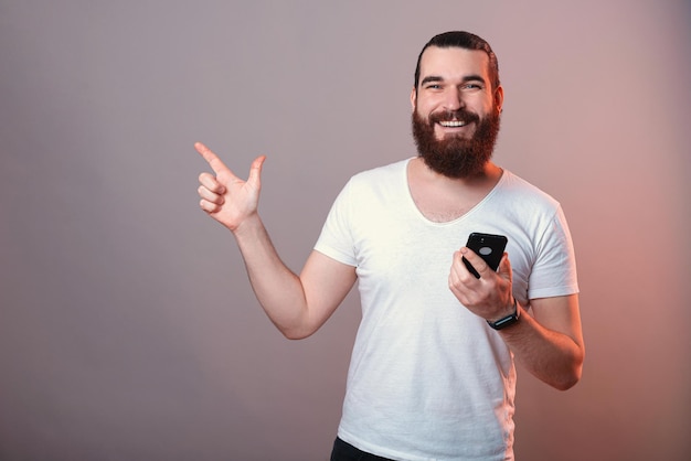 Ecstatic bearded man is pointing aside while holding his phone in hand