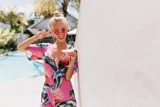 Ecstatic fairhaired girl with beautiful smile drinking cocktail Outdoor shot of slim inspired woman in pink attire spending time at resort