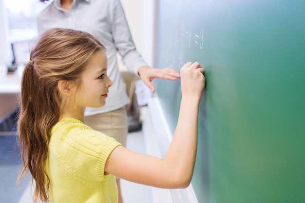 education, elementary school, learning, math and people concept - little smiling schoolgirl writing numbers on green chalk board in classroom