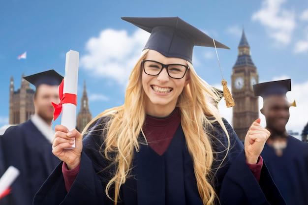 education, gesture and people concept - group of happy international students in mortarboards and bachelor gowns with diplomas celebrating successful graduation over london background