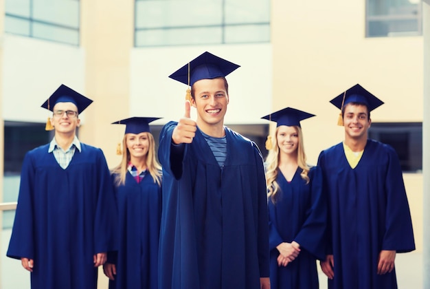 education, graduation, gesture and people concept - group of smiling students in mortarboards and gowns showing thumbs up outdoors