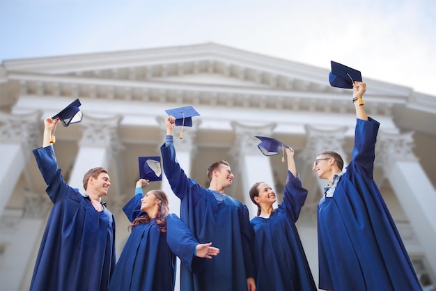 education, graduation and people concept - group of smiling university graduates in gowns waving mortarboards outdoors