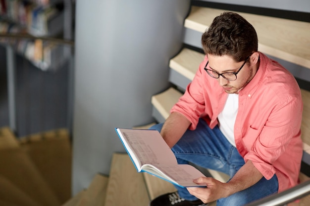 education, high school, university, learning and people concept - student boy or young man reading book sitting on stairs at library