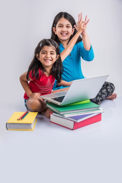 Education at home concept - 2 Cute little playful Indian or Asian girls studying or doing homework on laptop over pile of books while sitting on the floor at home. Isolated over white background