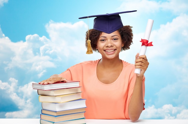 education, school, knowledge, graduation and people concept - happy smiling african american student girl in bachelor cap with books and diploma sitting at table over blue sky and clouds background