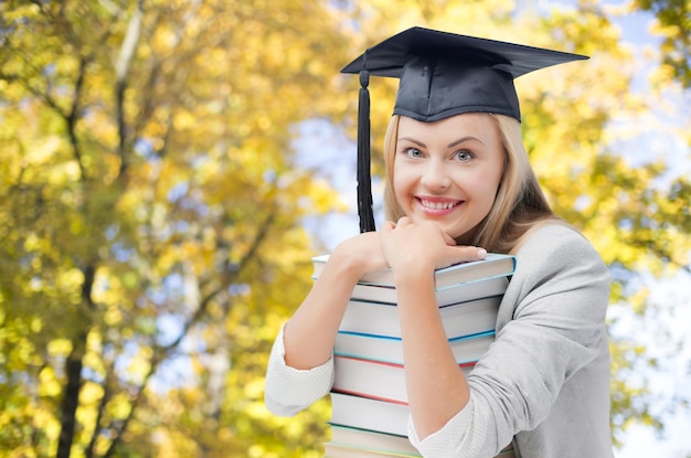 education, school, knowledge, graduation and people concept - happy smiling student girl in bachelor cap with books over autumn trees background