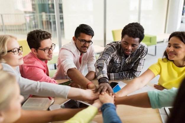 education, school, teamwork and people concept - group of international students with hands on top of each other sitting at table