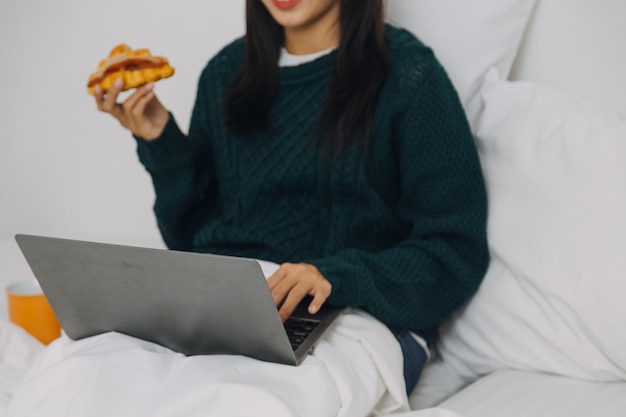 Efficient Young Millennial Girl Sitting on a Bed in the Morning Uses Laptop Computer and Eats Croissants and Drinks Coffee for Breakfast Top Down Shot