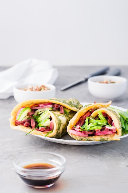 Egg rolls filled with pastrami, vegetables and green onions on a plate, sprouted grains and soy sauce in bowls on the table.