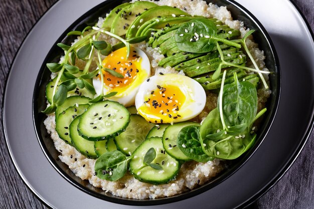 Egg Topped Quinoa Bowl with sunflower microgreens, cucumber, avocado, baby spinach in a black bowl on a dark wooden table, close-up, Turkish cuisine