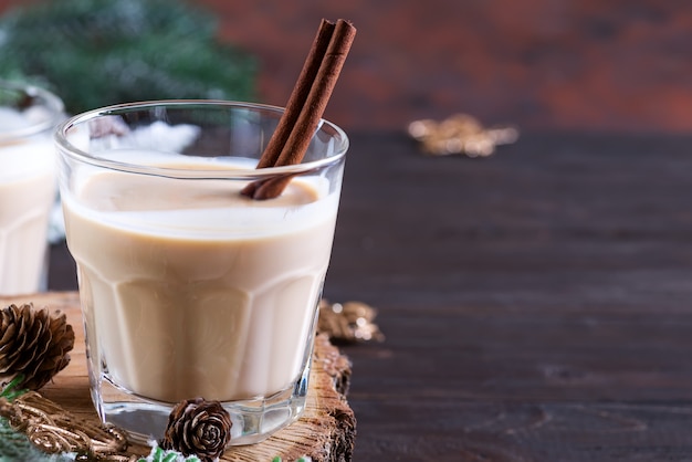 Eggnog Christmas milk cocktail with cinnamon, served in two glasses on a wood cutting board with fir branch and pine cone on dark wooden 