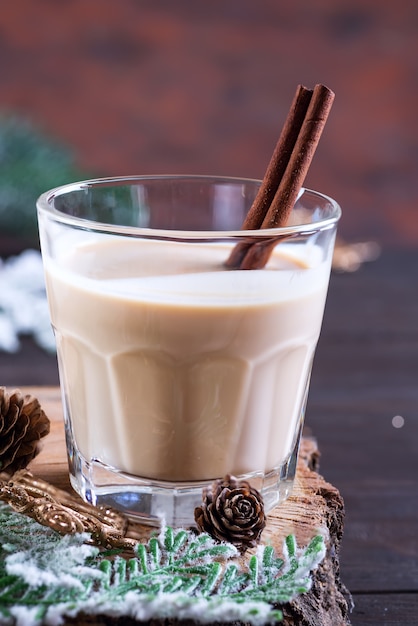 Eggnog Christmas milk cocktail with cinnamon, served in two glasses on a wood cutting board with fir branch and pine cone on dark wooden 