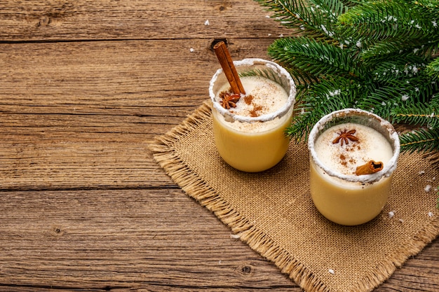 Eggnog cocktail in two glasses arranged with christmas decoration on old wooden table. Evergreen fer tree branch, artificial snow, sackcloth napkin