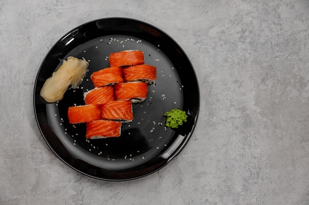 Photo eight servings of salmon and cucumber sushi with vossabi sauce and ginger on a black plate.