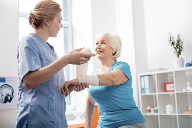 Elastic bandage. Pleasant delighted nurse looking at her patient while holding a white bandage