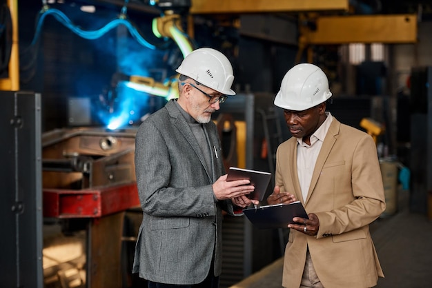 Elder businessman showing the results on tablet pc to his colleague while he filing the form during their work in plant