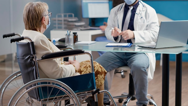 Elder patient with physical disability talking to doctor at checkup appointment in cabinet. Senior woman in wheelchair receiving medical treatment to help with rehabilitation.