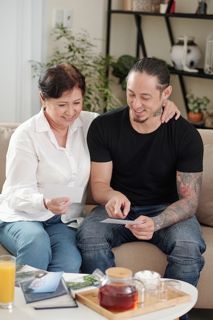 Elder son sitting together with his mother on the sofa and looking through the photos from album while they drinking tea