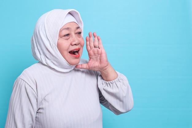 Elderly Asian Muslim woman wearing a headscarf shouting and screaming loud with a hand on her mouth