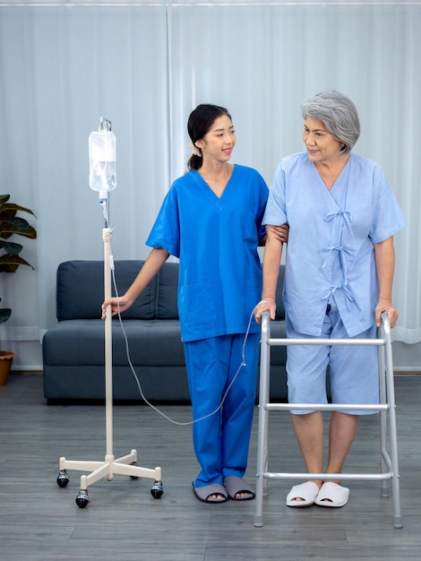 Elderly Asian woman patient trying to walk on walking frame held and carefully supported in arms by caregiver young polite female assistant nurse in blue suit in white room senior care concept