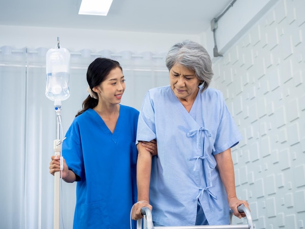 Elderly Asian woman patient trying to walk on walking frame held and carefully supported in arms by caregiver young polite female assistant nurse in blue suit in white room senior care concept