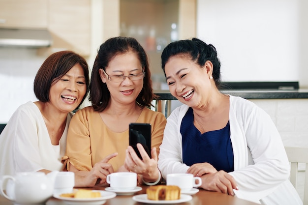 Elderly Asian woman showing funny photos on smartpone screen to her friends when they are sitting at kitchen table