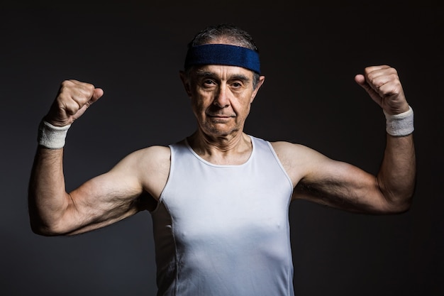 Photo elderly athlete wearing white sleeveless shirt, with sun marks on the arms, and blue headband, squeezing the biceps, on a dark background. sports and victory concept.