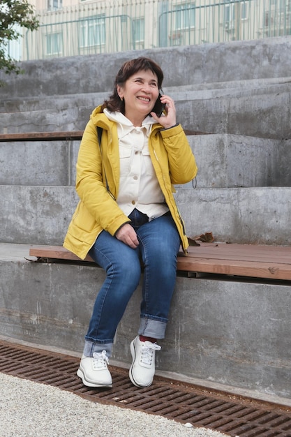 Elderly brunette woman sits in a city park on the steps smiling while talking on the phone