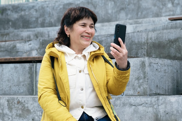 Elderly brunette woman sits in a city park on the steps smiling while talking on a video link