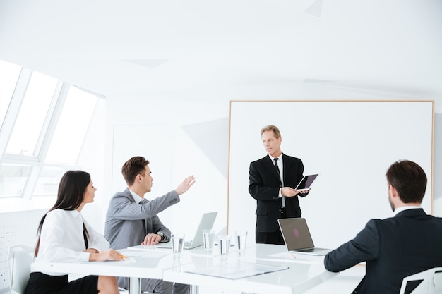 Elderly business man standing near the board and showing blank tablet computer screen to his colleagues on conference