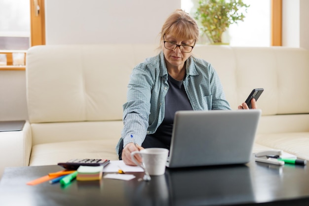 Elderly businesswoman working from home office talking on mobile phone call laughing enjoying telephone conversation using online app on laptop for consulting client