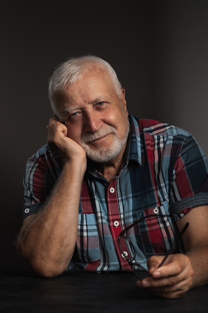 Elderly charismatic man propped his head on his hand, holds glasses in his hand and smiles while looking at the camera