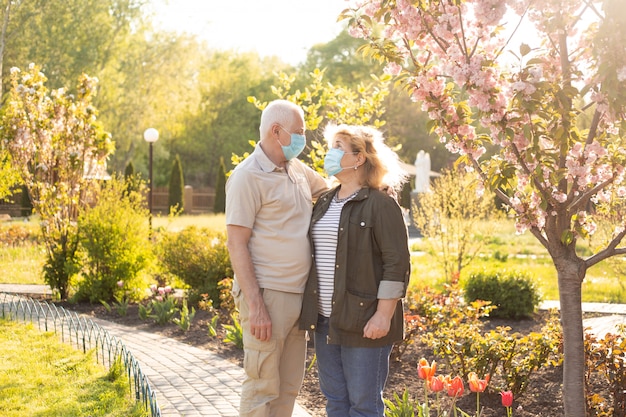 Elderly couple embracing in spring or summer park wearing medical mask to protect from coronavirus