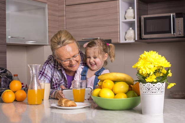 Elderly female and her adorable granddaughter watching a funny video together in a kitchen