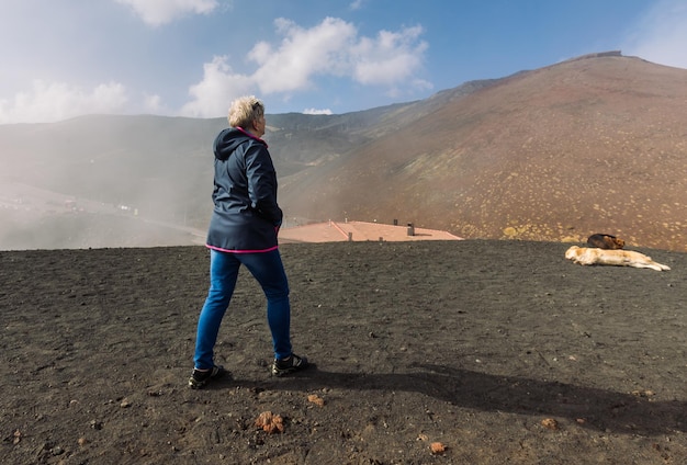 Elderly female traveler admiring breathtaking volcanic landscape of Etna covered with clouds at Catania Sicily, Italy.