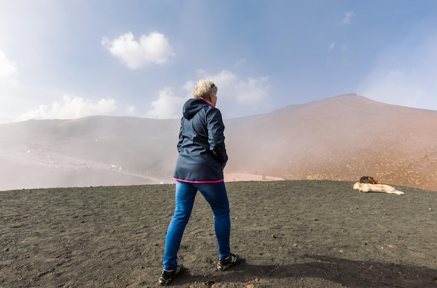 Elderly female traveler admiring breathtaking volcanic landscape of Etna covered with clouds at Catania Sicily, Italy.