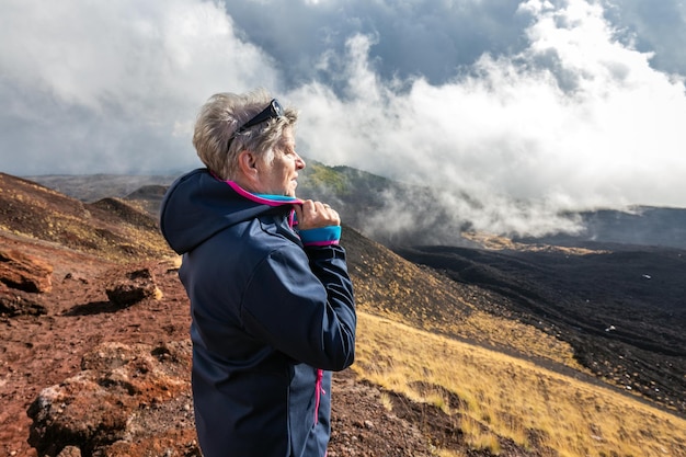 Elderly female traveler admiring breathtaking volcanic landscape of Etna covered with clouds at Catania Sicily, Italy.