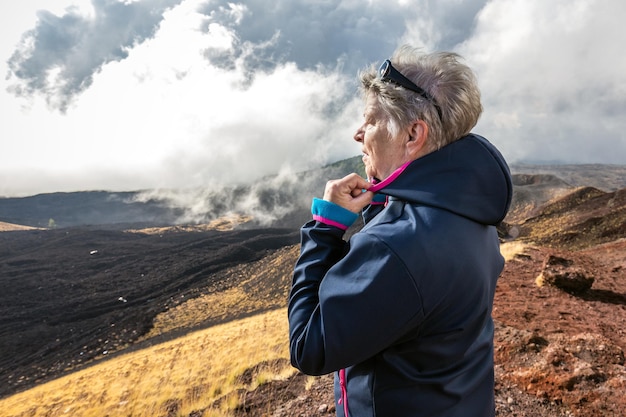 Elderly female traveler admiring breathtaking volcanic landscape of Etna covered with clouds at Catania Sicily, Italy.