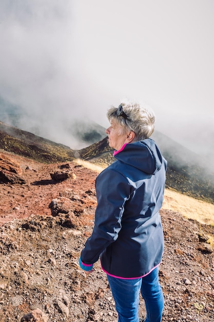 Elderly female traveler admiring breathtaking volcanic landscape of Etna covered with clouds at Catania Sicily, Italy.