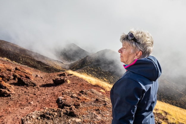 Elderly female traveler admiring breathtaking volcanic landscape of Etna covered with clouds at Catania Sicily, Italy.